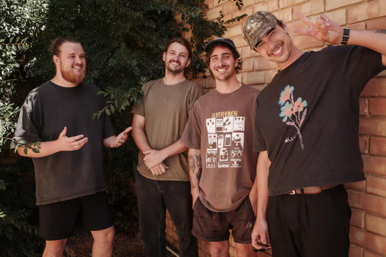 Four members of Australian indie rock band Sly Withers smiling together outdoors in front of a brick wall.